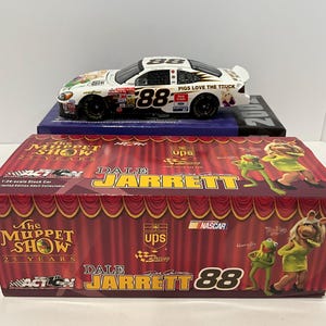 May include: A white and black Dale Jarrett #88 NASCAR stock car with "Pigs Love the Truck" text, displayed on a blue box. The car is above a box featuring Muppet Show characters and the text "Dale Jarrett 88".
