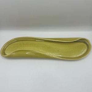 Puede incluir: Una fuente larga y curvada de cerámica en un tono amarillo verdoso apagado. La fuente tiene un acabado liso y brillante y un diseño ondulado único. Los bordes se curvan hacia arriba, creando un espacio poco profundo y abierto para servir alimentos o exhibir artículos.