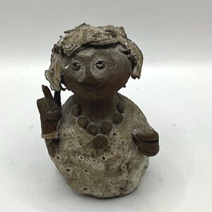 May include: A handcrafted ceramic figurine of a smiling person with a peace sign necklace and a raised index and middle finger. The figure has a textured, speckled surface with brown and beige tones.