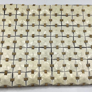 Könnte beinhalten: Rechteckige Clutch in Cremefarbe mit geometrischem Muster. Die Oberfläche besteht aus kleinen, quadratischen Elementen, die jeweils durch eine dunkle Linie verbunden sind. Kleine, rechteckige, braune Akzente sind auf jedem Quadrat sichtbar. Die Clutch besteht aus flexiblem Material.