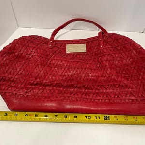 May include: A red handbag with a woven design and a top handle. The bag has a rectangular shape with a base that measures approximately 16 inches. A small gold-colored plaque is centered on the front of the bag.