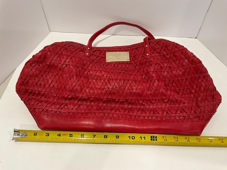 May include: A red handbag with a woven design and a top handle. The bag has a rectangular shape with a base that measures approximately 16 inches. A small gold-colored plaque is centered on the front of the bag.