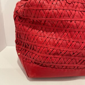 May include: A red leather tote bag with a woven design. The bag features a structured base and a soft, gathered top. Gold-colored studs are visible on the bag's exterior. The bag is made of a textured leather material.
