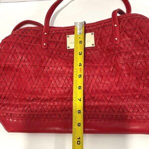 May include: Red handbag with woven texture and dual handles. The bag has a rectangular shape and measures approximately 10 inches in height. Gold-toned metal accents and a rectangular nameplate are visible on the front.