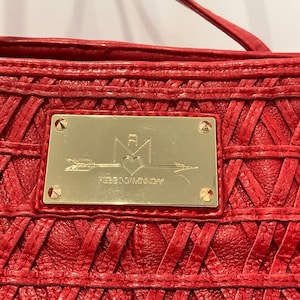 May include: Close-up of a red leather handbag with a woven design. A gold-colored rectangular plaque is centered on the bag, featuring a logo with an arrow, heart, and the brand name "REBECCAMINKOFF". The bag has a top handle.