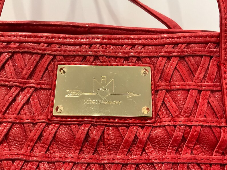 May include: Close-up of a red leather handbag with a woven design. A gold-colored rectangular plaque is centered on the bag, featuring a logo with an arrow, heart, and the brand name "REBECCAMINKOFF". The bag has a top handle.