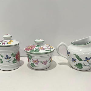 May include: A white ceramic kitchen set featuring floral designs. The set includes a lidded canister, a lidded bowl, and a creamer pitcher. The floral pattern includes pink, yellow, and purple flowers with green leaves.