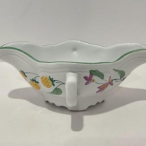 May include: White ceramic gravy boat with a scalloped rim and a handle. The boat is decorated with yellow and green fruit and purple flowers. A thin green line trims the rim.