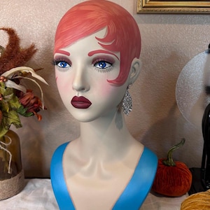Puede incluir: Cabeza de maniquí decorativa con un estilo art déco vintage. La cabeza presenta una tez pálida, cabello rojo, ojos azules y labios rojo oscuro. El maniquí lleva una blusa azul y pendientes plateados. La estética general recuerda a la década de 1920.
