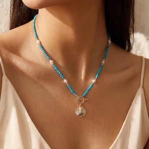 May include: A necklace featuring small turquoise beads interspersed with white pearls, finished with a gold-toned toggle clasp and a gold-toned seashell pendant. The jewelry piece rests against the neck, showcasing a vibrant blue and white color palette.