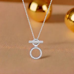 May include: A silver necklace featuring a circular pendant and a horizontal bar, both adorned with small, clear gemstones. The necklace is suspended from a fine chain, set against a warm, neutral backdrop.