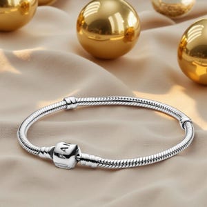 May include: A silver bracelet featuring a snake chain design and a square clasp. The clasp is engraved with the letters "PAN". The bracelet is presented on a beige surface, with golden spheres visible in the background.