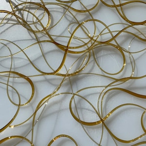 May include: A close-up of a white surface with a pattern of gold, metallic, interwoven, wavy lines.