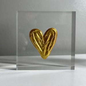 May include: A golden heart sculpture encased in a clear, square acrylic block. The heart has a textured surface and is centered within the block. The background is a gradient of gray and white.