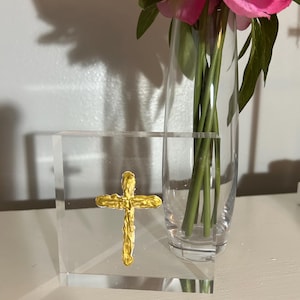 May include: A gold cross is displayed on a clear acrylic block. The block is square shaped and has beveled edges. The cross is textured and has a raised design. The block is sitting on a white surface.