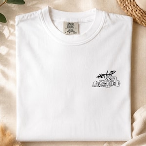 May include: A white short-sleeved t-shirt featuring a black line-art graphic of a race car on the right chest area. The text "HOT LAP" is printed in a stylised black font above the car. The inner neck label reads "COMFORT COLORS" and "P. CH".