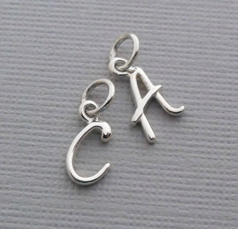 K&ouml;nnte beinhalten: Zwei silberne Anh&auml;nger in Form der Buchstaben 'A' und 'C'. Die Buchstaben sind in einer kursiv geschriebenen Schriftart.