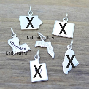 May include: A set of six silver state charm pendants with a black letter "X" on each. The charms are shaped like the states of Florida, Louisiana, Iowa, Utah, Wyoming, and Maine. The charms are labeled with the state name in a dark font. The charms are labeled "Natural Letters" and "Dark Letters".