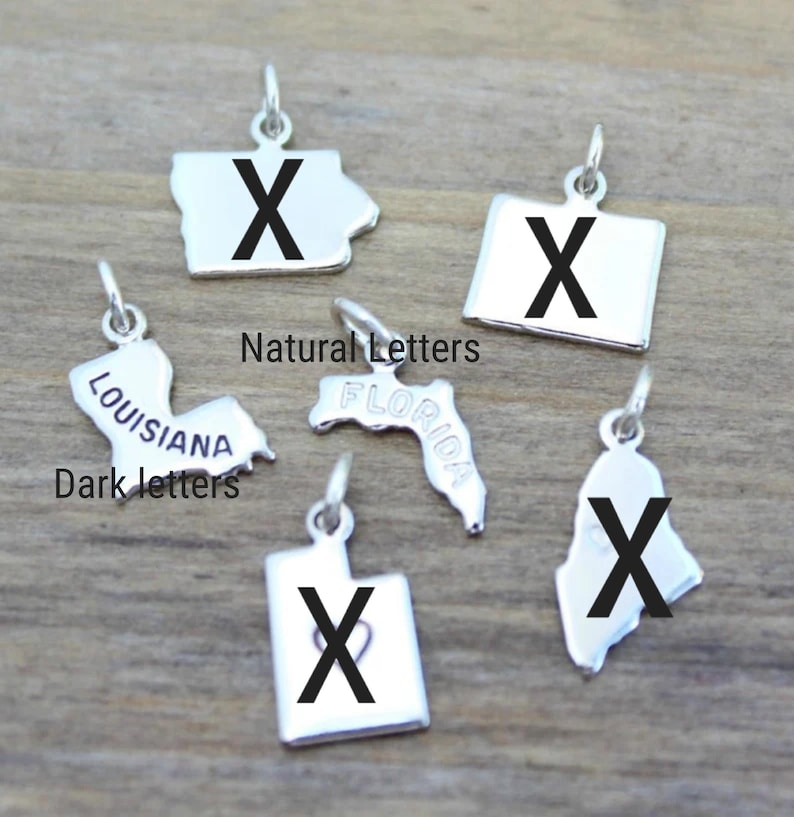 May include: A set of six silver state charm pendants with a black letter "X" on each. The charms are shaped like the states of Florida, Louisiana, Iowa, Utah, Wyoming, and Maine. The charms are labeled with the state name in a dark font. The charms are labeled "Natural Letters" and "Dark Letters".