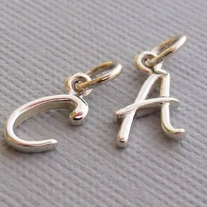 K&ouml;nnte beinhalten: Zwei silberne Anh&auml;nger in Form der Buchstaben 'C' und 'A'. Die Buchstaben sind in einer kursiv geschriebenen Schriftart.