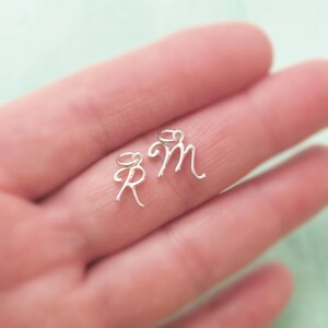 K&ouml;nnte beinhalten: Zwei silberne Anh&auml;nger in Form der Buchstaben R und M. Die Anh&auml;nger werden auf einer Hand gezeigt.