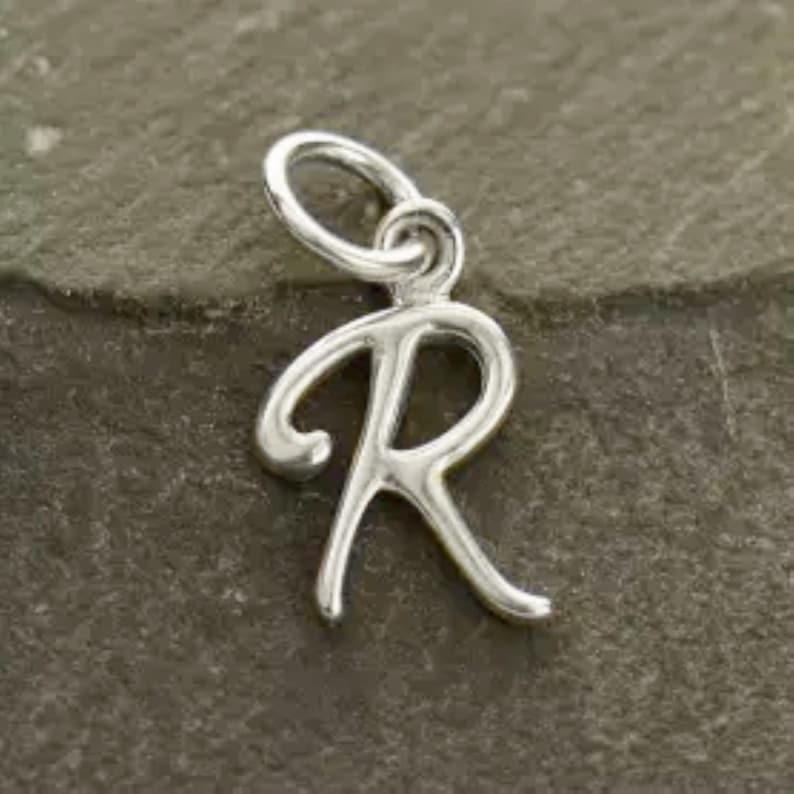 K&ouml;nnte beinhalten: Silberner Anh&auml;nger in Form des Buchstabens R. Der Buchstabe ist in einer kursiven Schriftart gestaltet.