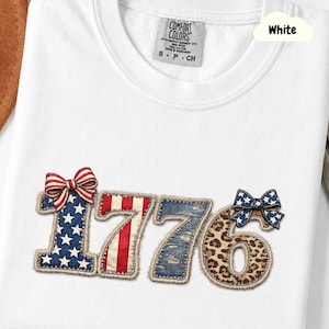 Peut inclure: T-shirt blanc avec le chiffre "1776" appliqué sur le devant. Les chiffres sont décorés de motifs patriotiques, dont des étoiles et des rayures, du denim et un imprimé léopard. Un nœud rouge, blanc et bleu est sur le dessus du "1" et du "6".