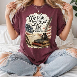 May include: Burgundy t-shirt featuring a graphic with the text "We the People" and "250 Years of Freedom". The graphic includes an American flag, a quill, inkwell, and books. The shirt is a short-sleeved, crew neck style.