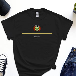 Puede incluir: Camiseta negra con una franja roja, amarilla y verde a través del pecho. La camiseta presenta el escudo de armas de Bolivia y la palabra "Bolivia" impresa debajo de la franja.