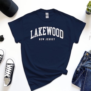 May include: Navy blue t-shirt with the white text "Lakewood New Jersey". The shirt is surrounded by black sunglasses, black sneakers, a black cap, a succulent plant, a smartwatch, and blue jeans.