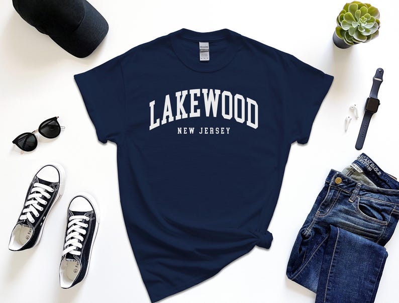 May include: Navy blue t-shirt with the white text "Lakewood New Jersey". The shirt is surrounded by black sunglasses, black sneakers, a black cap, a succulent plant, a smartwatch, and blue jeans.