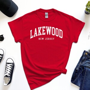 May include: A red t-shirt with the white text "Lakewood New Jersey" in an arched font. The shirt is surrounded by black sunglasses, black sneakers, a black cap, a watch, and blue jeans.