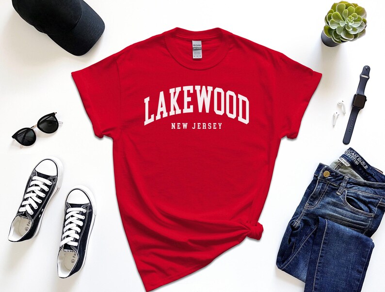 May include: A red t-shirt with the white text "Lakewood New Jersey" in an arched font. The shirt is surrounded by black sunglasses, black sneakers, a black cap, a watch, and blue jeans.
