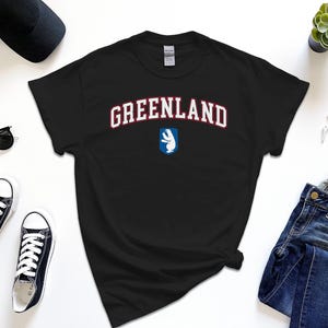 Op de afbeelding: Zwart T-shirt met witte tekst die "GREENLAND" in een college-lettertype leest. Een witte ijsbeergrafiek bevindt zich onder de tekst.
