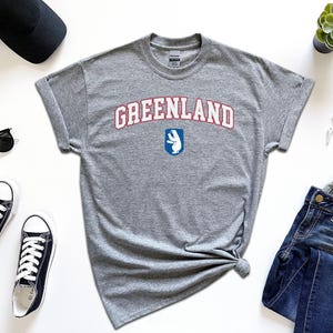 Op de afbeelding: Grijs T-shirt met de tekst "GREENLAND" in rode en blauwe letters met een witte ijsbeer afbeelding.