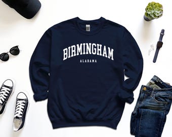以下が含まれることがあります： 紺色のスウェットシャツに、白い文字で「BIRMINGHAM ALABAMA」と書かれています。 サングラス、スニーカー、腕時計などのアクセサリーも写っています。 カジュアルな服装です。