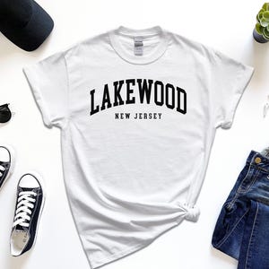 May include: A white t-shirt with the text "Lakewood New Jersey" in black, paired with black sunglasses, sneakers, a black cap, a smartwatch, and blue jeans. A small succulent plant is also visible.