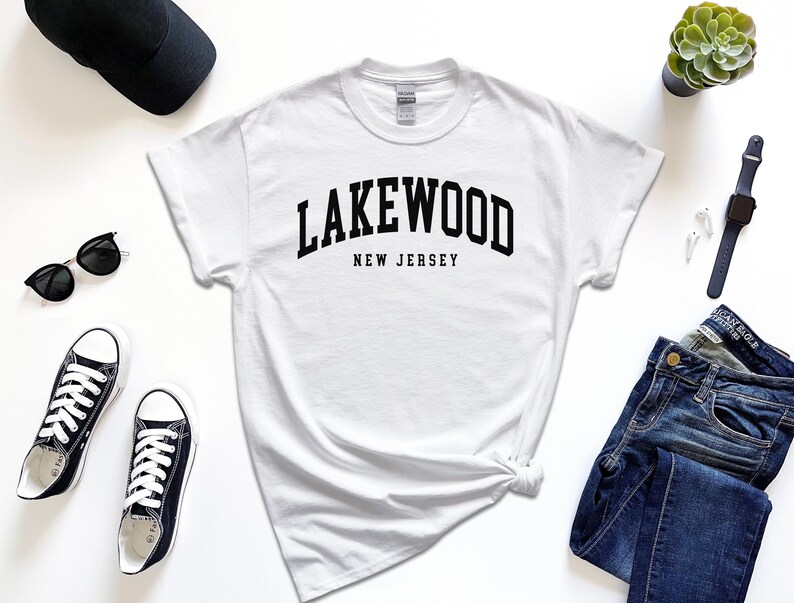 May include: A white t-shirt with the text "Lakewood New Jersey" in black, paired with black sunglasses, sneakers, a black cap, a smartwatch, and blue jeans. A small succulent plant is also visible.