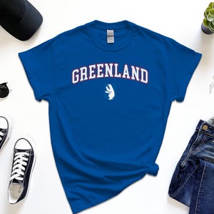 Op de afbeelding: Een koninklijk blauw t-shirt met een witte ijsbeer afbeelding en de tekst "GREENLAND" in wit met rode contouren.