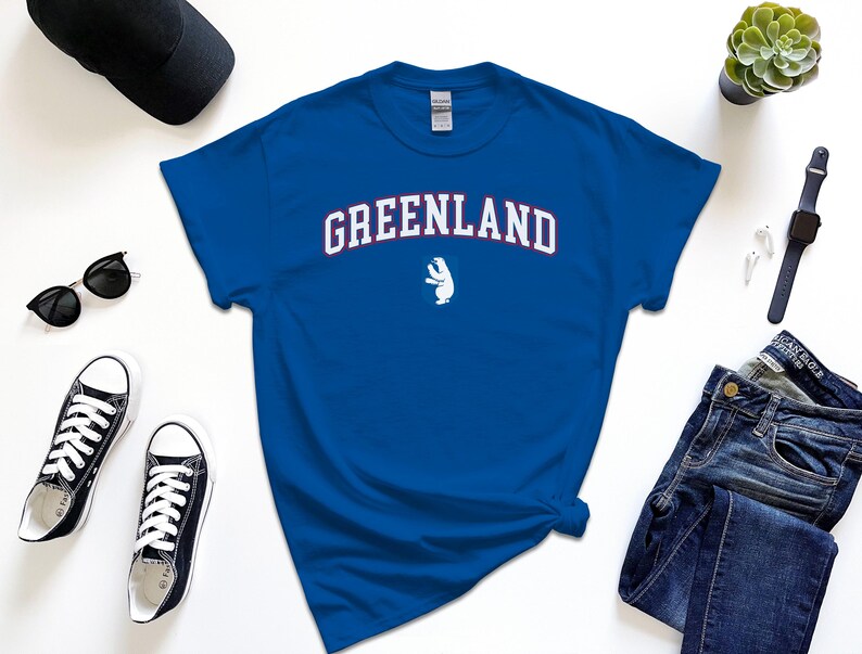 Op de afbeelding: Een koninklijk blauw t-shirt met een witte ijsbeer afbeelding en de tekst "GREENLAND" in wit met rode contouren.