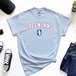 Op de afbeelding: Lichtblauw T-shirt met de tekst "GREENLAND" in rood en witte letters met een wit ijsbeerlogo in een blauw schild.