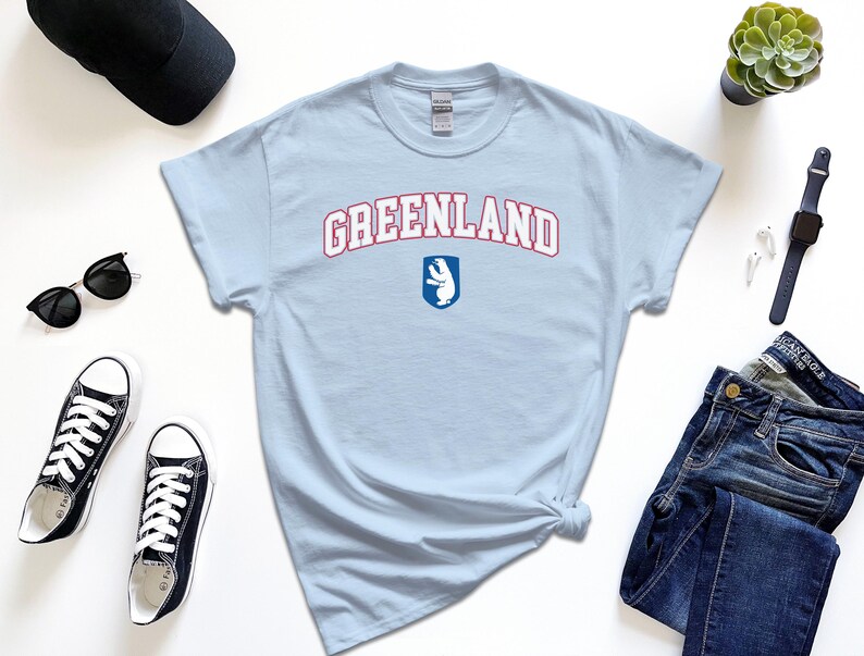 Op de afbeelding: Lichtblauw T-shirt met de tekst "GREENLAND" in rood en witte letters met een wit ijsbeerlogo in een blauw schild.