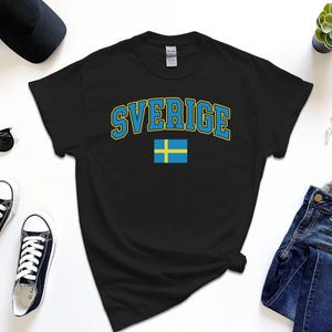 May include: Black t-shirt with the word "SVERIGE" in blue and yellow lettering, and a small Swedish flag. The shirt is laid flat on a white surface, surrounded by accessories like sunglasses, sneakers, and a watch.