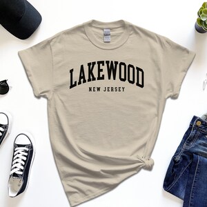 May include: A beige t-shirt with the word "Lakewood" arched across the chest in black, with "New Jersey" below. The shirt is styled with black sunglasses, sneakers, a watch, a cap, and jeans.