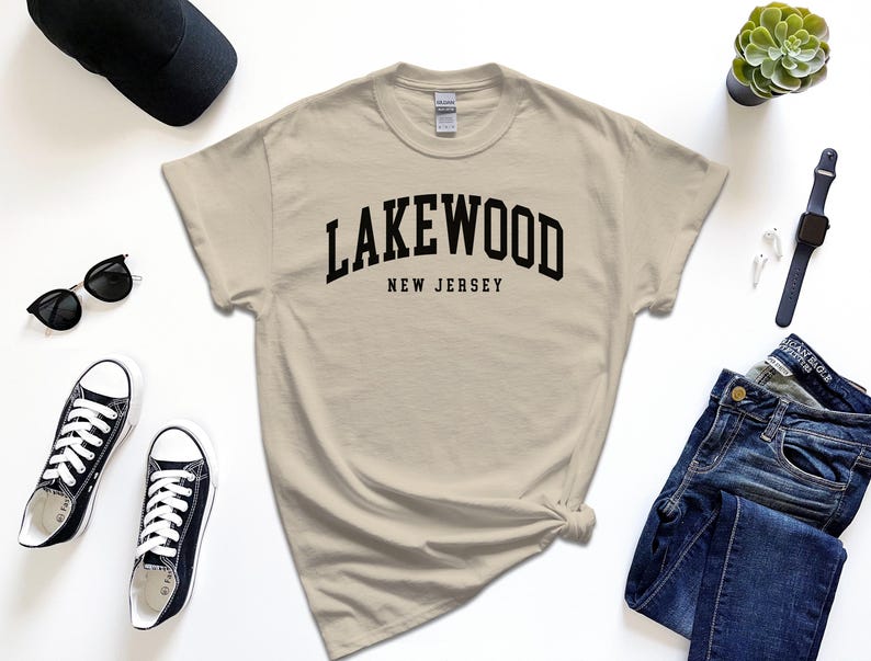 May include: A beige t-shirt with the word "Lakewood" arched across the chest in black, with "New Jersey" below. The shirt is styled with black sunglasses, sneakers, a watch, a cap, and jeans.
