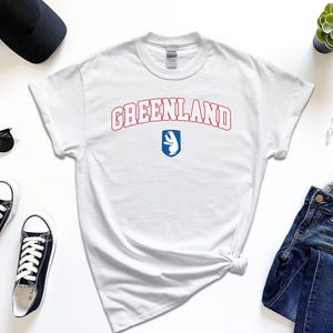 Op de afbeelding: Wit T-shirt met een rode en blauwe grafische afbeelding met de tekst "GREENLAND" en een ijsbeerlogo.