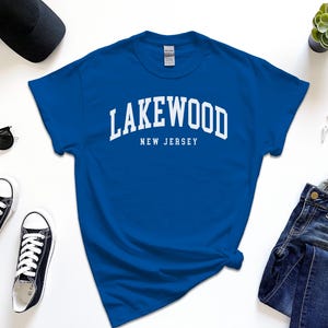 May include: Royal blue t-shirt with the word "Lakewood" arched across the chest in white block letters, with "New Jersey" below. The shirt is surrounded by black sunglasses, black sneakers, a black cap, a watch, and jeans.