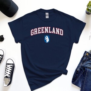 Op de afbeelding: Een marineblauw t-shirt met het woord "GREENLAND" in wit met rode omlijsting. Een witte ijsbeer bevindt zich in een blauw schild onder de tekst.
