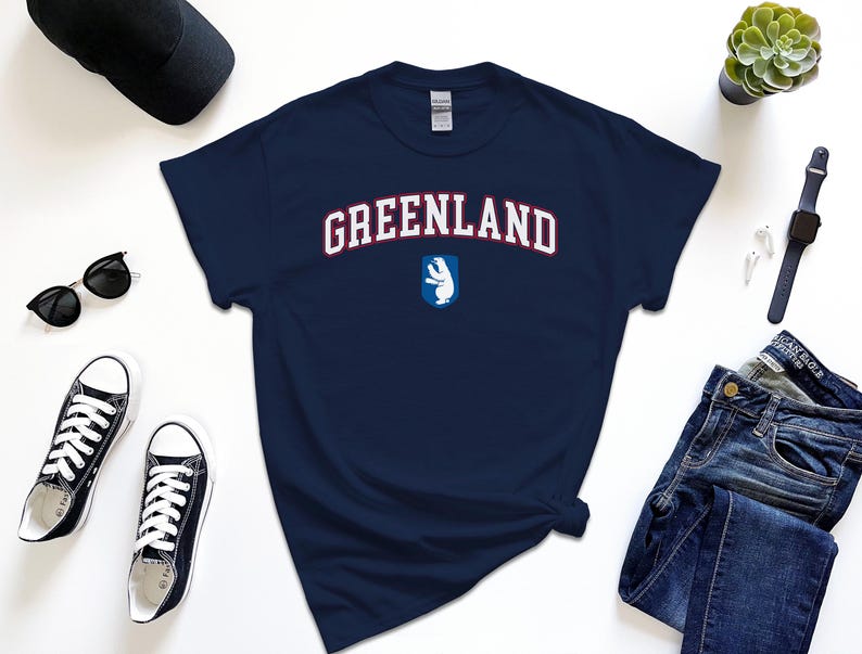 Op de afbeelding: Een marineblauw t-shirt met het woord "GREENLAND" in wit met rode omlijsting. Een witte ijsbeer bevindt zich in een blauw schild onder de tekst.