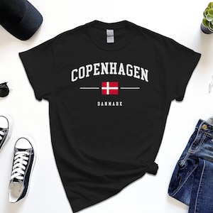 May include: Black t-shirt with white text that reads "Copenhagen" with a line above and below the text. The Danish flag is centered between the lines. Below the flag is the word "Danmark" in white text.